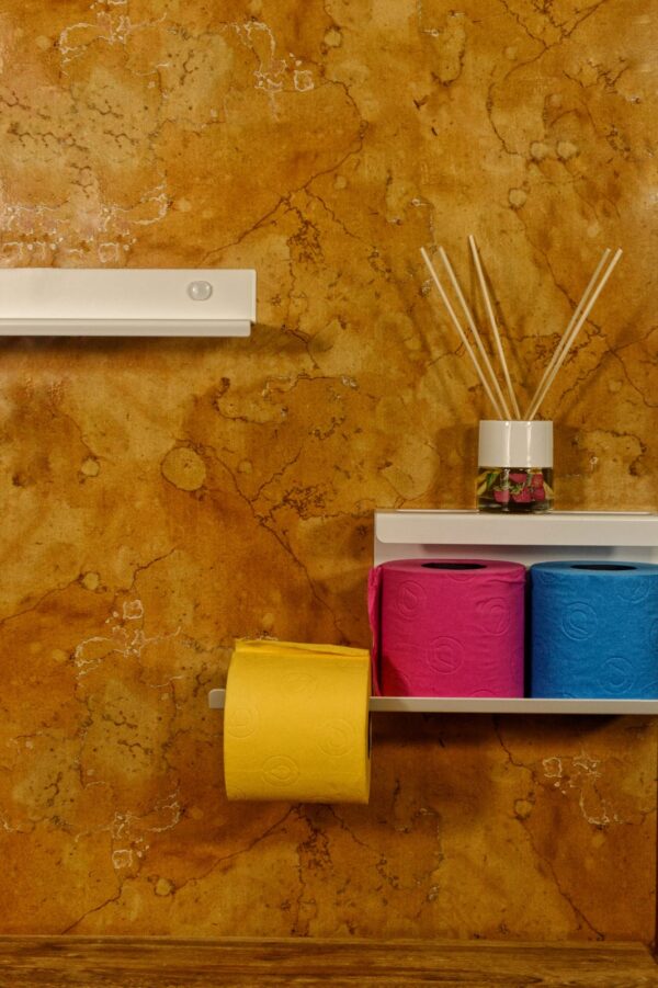 Una revista de estilo de vida, un difusor de aroma y un rollo de papel higiénico de color rosa brillante descansan en un estante de pared minimalista contra una pared de textura terrosa.