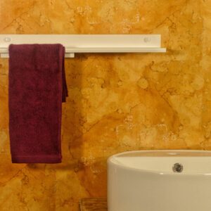 La imagen presenta un primer plano de un lavabo de baño moderno. El lavabo es blanco, con forma ovalada y está colocado sobre un mueble pequeño de madera de color marrón claro. La madera parece de una textura rugosa y ligeramente desgastada, lo que le da una calidez rústica. La pared detrás del lavabo es la característica más llamativa de la imagen: es de un color dorado-marrón con una textura marmórea irregular, que se asemeja a una pared enlucida o con un efecto estuco. El color dorado crea una sensación cálida y un tanto opulenta. Hay una textura irregular con vetas y manchas, que le dan una apariencia natural y orgánica. En la pared, a una altura media, se encuentra una repisa horizontal blanca y estrecha. Sobre la repisa cuelga una toalla de color borgoña oscuro. La toalla es suave y está doblada sencillamente, colgando con naturalidad sobre la repisa. El color contrastante de la toalla, contra la pared dorada y la repisa blanca, añade interés visual. La atmósfera de la imagen es serena, limpia y relajante. Los colores cálidos y la apariencia de los materiales crean una sensación acogedora y de lujo discreto. El estado de ánimo es tranquilo y silencioso, propio de un espacio de baño privado. La composición es simple y minimalista, enfocándose en los elementos de diseño del espacio.